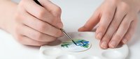 Young woman with fair skin mixing blue and green watercolor paint on white ceramic palette using thin brush for artistic creation.