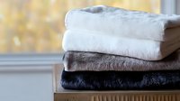 Neatly folded stack of soft plush blankets in white, gray and black colors on wooden surface near window, cozy home textile arrangement for interior decor.