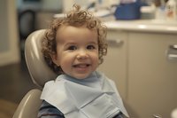 Smiling curly-haired toddler wearing light blue dental bib sits in dental chair during routine checkup, showing positive pediatric dentistry experience.