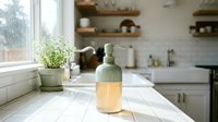 Green glass soap dispenser with wooden base on white kitchen counter near window with herb plant creating clean modern home interior atmosphere.