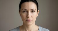 Serious middle-aged Caucasian woman with dark hair pulled back looking directly at camera against neutral background for professional headshot.