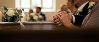 Elderly hands clasped in prayer during funeral service with white roses and mourners in church pew, expressing grief, remembrance and spiritual comfort.