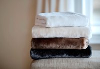 Stack of folded soft plush blankets in white, brown and black colors on beige sofa, cozy home textile arrangement with natural daylight from window.