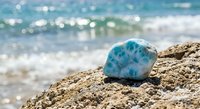 Larimar gemstone on rocky shore with turquoise ocean waves in background. Natural blue stone mineral specimen for jewelry making and crystal healing practices.