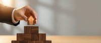 Hand placing wooden house block on top of stacked wooden blocks, representing real estate investment, property development, and home building concepts for business and finance.