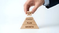 Male hand placing wooden cube on pyramid stack with strategy, plan, and growth text layers, representing business development and organizational hierarchy.