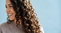 Young woman with curly brown hair smiling outdoors against blue sky background. Natural beauty portrait for lifestyle and wellness concepts.