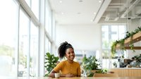 Young woman with dark skin tone and black hair wearing a mustard yellow shirt, smiling while working at a modern office reception desk with plants and natural light from large windows.
