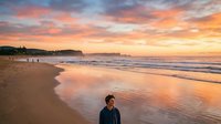 Man in dark jacket standing alone on sandy beach at sunset with golden and pink sky, gentle waves, and distant cliffs, suitable for travel and lifestyle content.