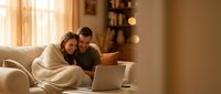 Young couple relaxing on couch with laptop in cozy living room during golden hour. Intimate moment of togetherness and modern lifestyle at home.