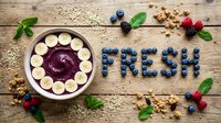 Acai smoothie bowl with banana slices on rustic wood, surrounded by blueberries spelling FRESH, granola, raspberries, and mint leaves.