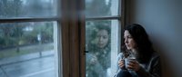 Young woman with long dark hair and fair complexion, wearing casual clothing, holding a warm beverage while gazing thoughtfully out a window on a rainy day. Suitable for lifestyle, wellness, and seasonal content.