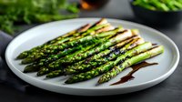 Grilled green asparagus spears on white ceramic plate drizzled with balsamic reduction sauce, close-up view showing charred marks and fresh preparation.