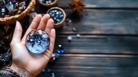 Female hand holding crystal sphere with blue reflections against rustic wooden background with scattered gems and star anise, creating mystical atmosphere.