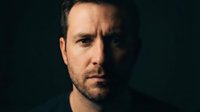 Serious Caucasian man with beard looking directly at camera with intense expression against dark background for dramatic portrait photography.