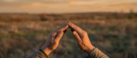 Hands forming protective shelter gesture over rural landscape at golden hour sunset, symbolizing care, protection, and environmental conservation concepts.