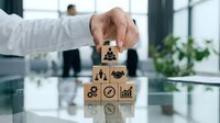 Male hand placing wooden block with people icon on stack of business symbols in modern office setting with colleagues and plants visible in background.