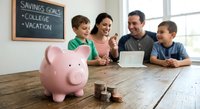Happy family of four planning finances with pink piggy bank and coins on wooden table discussing college and vacation savings goals together.