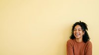 Young woman with curly dark hair and medium skin tone wearing a rust-colored ribbed sweater, smiling against a soft yellow background, suitable for lifestyle and portrait projects.