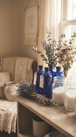 Cozy farmhouse interior with blue glass bottles, dried lavender, eucalyptus and sea salt jars on rustic wooden table creating peaceful spa atmosphere.