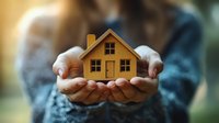 Cupped hands gently holding small wooden house model against blurred green background, symbolizing property care, home ownership and real estate investment.