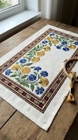 Traditional embroidered linen table runner with floral pattern featuring yellow marigolds and blue cornflowers on wooden table near window.