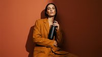 Professional businesswoman in orange suit holding black bottle against warm gradient background for corporate branding and product presentation.