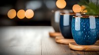Ceramic mugs with blue glaze sit on wooden coasters beside evergreen sprigs, warm bokeh lights glowing softly in background, creating cozy winter holiday atmosphere for seasonal beverage and home decor designs.