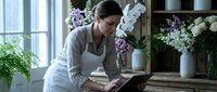 Young Caucasian female florist in apron working on tablet in flower shop surrounded by fresh blooms and arrangements for small business management.