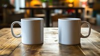 Two white ceramic coffee mugs sitting on rustic wooden table in cozy cafe setting with warm blurred background lighting for morning beverage concepts.