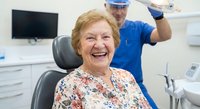 Happy senior Caucasian woman patient smiling in dental chair during checkup with male dentist in blue scrubs working behind her in modern clinic office.