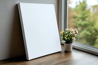 Blank white canvas mockup on wooden windowsill with small potted flowers and natural daylight, ready for artwork presentation or home decor display.