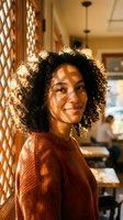 Young mixed-race woman with curly hair smiling warmly in cozy restaurant setting wearing rust-colored sweater with natural sunlight creating golden hour ambiance.