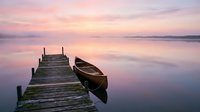 Wooden dock extending into calm lake at sunrise with moored boat, reflecting pink and purple sky. Serene landscape for travel, nature, and meditation projects.