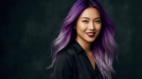 Young Asian woman with vibrant purple hair smiling confidently in black blazer against dark background for professional business portrait.