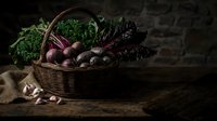 Wicker basket filled with fresh organic vegetables including kale, beets, and root vegetables on rustic wooden surface with burlap cloth for farm-to-table cooking and healthy eating concepts.