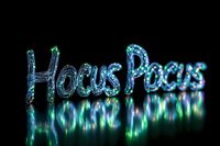 Glowing blue and green neon text spelling Hocus Pocus with magical sparkle effect and reflection on dark background, creating mystical Halloween atmosphere.