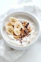 Fresh sliced bananas in creamy yogurt topped with dark chocolate shavings and caramel drizzle on white ceramic bowl, bright minimalist breakfast setup.