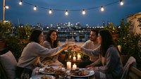 Four young women friends enjoying evening dinner party on rooftop terrace with string lights, toasting with wine glasses over city skyline at dusk.