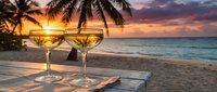 Two glasses of white wine on wooden table at tropical beach sunset with palm trees and ocean waves creating romantic vacation atmosphere.
