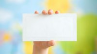 Caucasian hand holding blank white card outdoors with colorful Easter eggs on top and spring garden background, suitable for business cards, invitations, and promotional messaging.