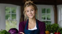 Young woman in kitchen with fresh vegetables, wearing burgundy cardigan and blue apron, smiling confidently while preparing healthy meal.