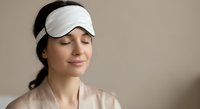 Young woman wearing white sleep mask with peaceful expression relaxing with closed eyes. Self-care wellness routine for better rest and meditation.