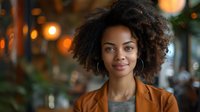 Beautiful young African American woman with natural curly hair smiling confidently, wearing brown blazer against blurred warm bokeh background, perfect for business or lifestyle content.