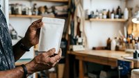 Artisan holding blank white kraft paper pouch in workshop studio with shelves of bottles and supplies, showcasing packaging for food, coffee, tea, or retail products.