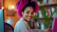 Young African American woman with vibrant pink curly hair smiling while working on laptop at home office, wearing white blouse and gold hoop earrings against blurred bookshelf background.