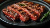 Juicy grilled beef steak sliced on black plate with rosemary sprig and seasonings. Medium rare meat with perfect grill marks shows pink center and charred crust.