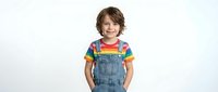Happy young boy with brown hair wearing rainbow striped shirt and denim overalls standing with hands in pockets against white background for children fashion.