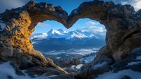Natural heart-shaped rock arch frames snow-capped mountain peaks at sunrise, dramatic winter landscape with golden morning light illuminating rugged stone formation.