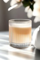 Ribbed glass with creamy coffee drink casting soft shadows on white surface, natural light streaming through floral background creates elegant mood.